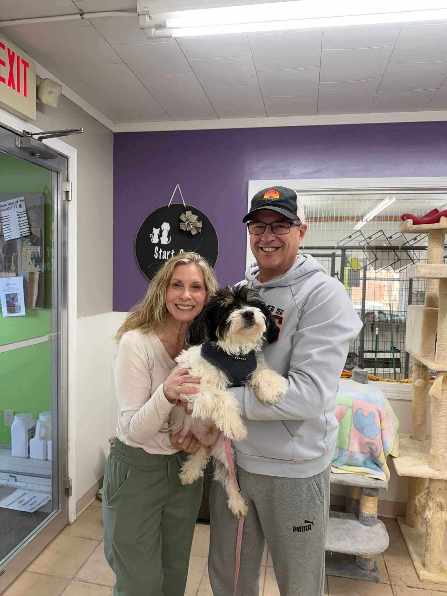 Two smiling adults holding a small black-and-white dog indoors