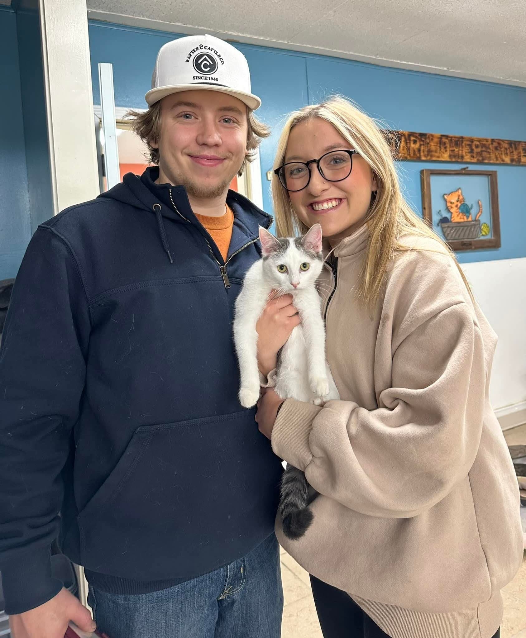 Two smiling adults pose indoors while one holds a white-and-gray cat